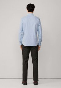 Camicia azzurra leggera, dalla vestibilità aderente, con maniche lunghe e polsini. Pantaloni scuri a fantasia e scarpe marroni, tutto visto da dietro.