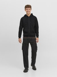 Juodas zip-up bliuzonas su raišteliais, derinamas su juodomis cargo kelnėmis, turinčiomis šonines kišenes, ir juoda avalyne. Neutralus fonas.