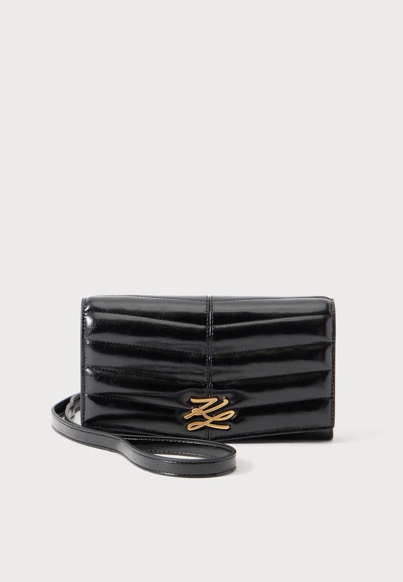 Pochette en cuir noir brillant matelassé avec logo décoratif doré et fine bride poignet attachée sur un fond clair.