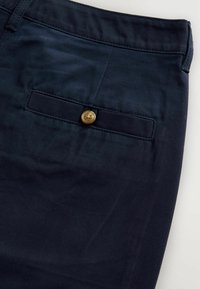 Pantalons bleu foncé avec une texture lisse, comportant une poche arrière avec un bouton beige. Le tissu présente un léger éclat.