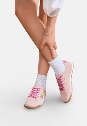 Jambes croisées portant des chaussettes blanches et des baskets roses avec des lacets roses et une étoile dorée, main avec bracelet en argent et bagues reposant sur le tibia.