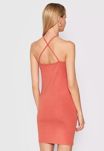 Donna con lunghi capelli biondi che indossa un vestito aderente senza maniche di colore corallo con sottili spalline incrociate, girata di spalle che mostra tatuaggi sulla schiena.