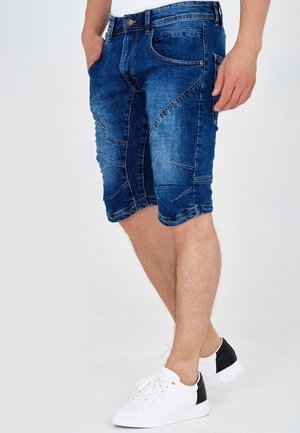 Man draagt donkerblauwe denim knielange shorts en witte sneakers met zwarte hielstukken, staat met één hand in de zak.