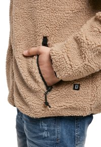 Chaqueta de forro polar beige con una superficie texturizada, que cuenta con un bolsillo lateral con una cremallera negra y un pequeño parche de logo en el bajo.