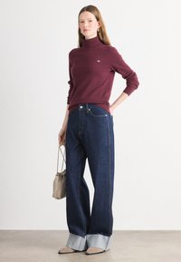 Burgunderfarbener Rollkragenpullover kombiniert mit weit geschnittenen, dunklen Jeans aus Denim, die ausgefranste, helle Denim-Bündchen haben. Das Modell hält eine kleine beige Handtasche.