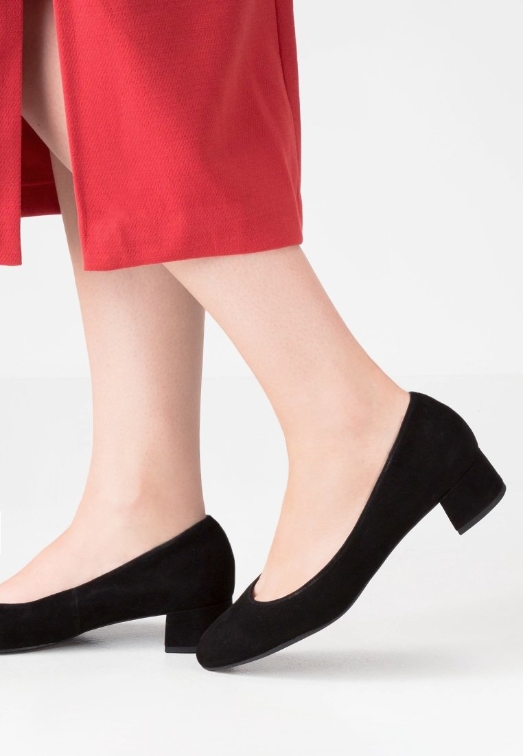 Femme portant des chaussures en daim noire à talons bas et une jupe rouge, debout sur un sol blanc avec un talon légèrement soulevé.