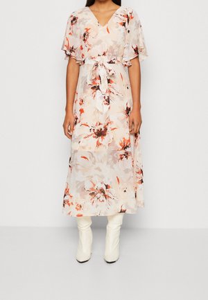 Femme portant une robe midi florale de couleur claire avec des manches volantées et une ceinture à la taille, associée à des bottes blanches hauteur genoux.