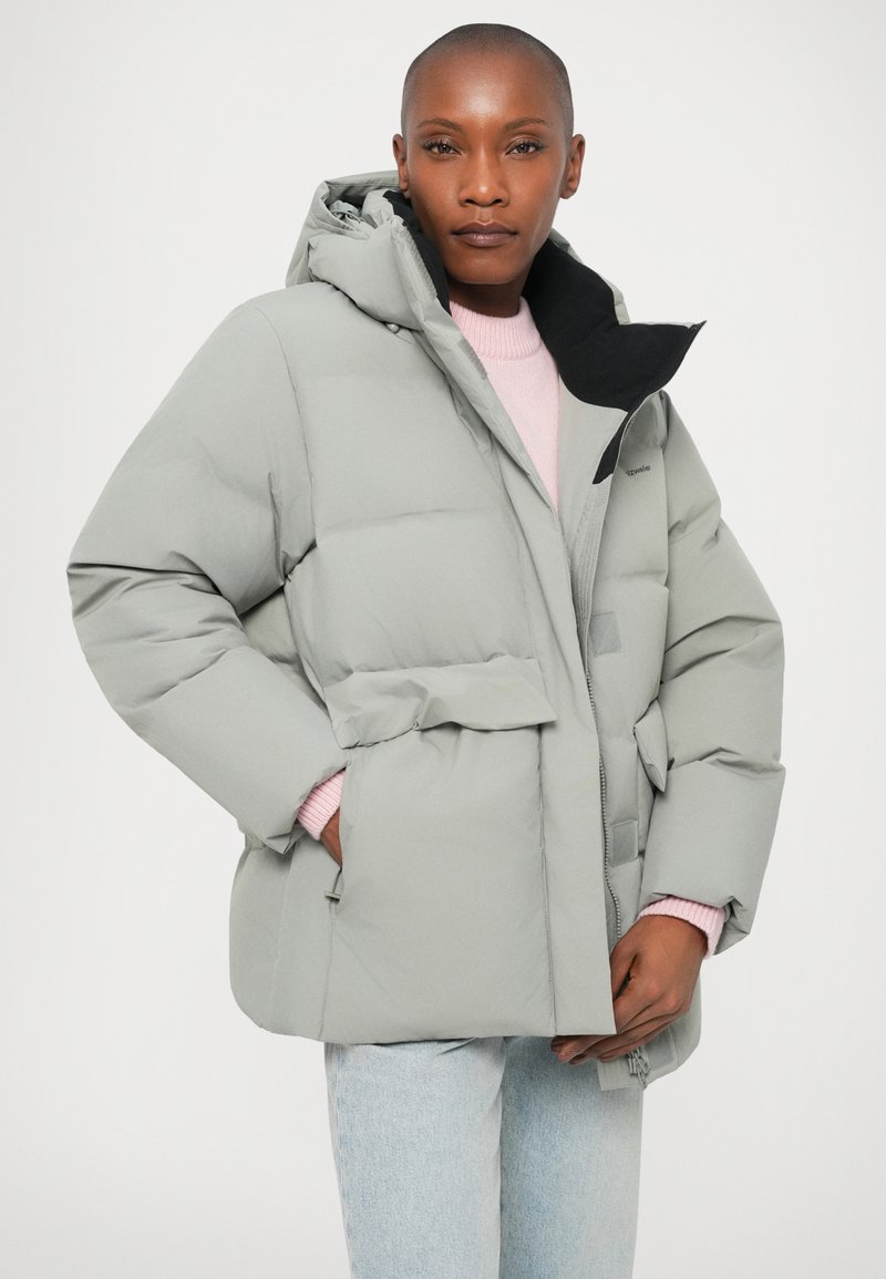 Veste grise matelassée avec capuche, fermeture éclair à l'avant et poches. Doublée de matériau noir, portée sur un pull rose clair. Coupe décontractée et texture lisse.