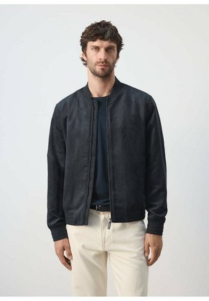 Mann mit kurzen lockigen Haaren, der eine schwarze Reißverschlussjacke, ein dunkles Hemd und hellbeige Hosen trägt und vor einem einfarbigen weißen Hintergrund steht.