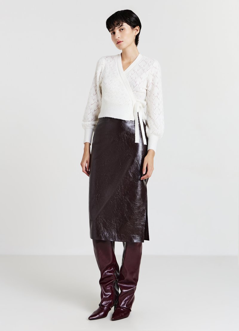 Maglione avvolgente a maglia color crema con scollo a V e motivo a rilievo, abbinato a una gonna midi in pelle lucida marrone e stivali al ginocchio coordinati.