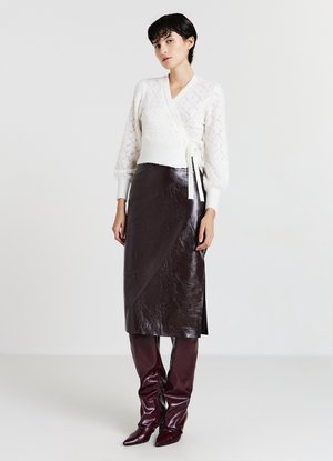 Cremfarbener gestrickter Wickelpullover mit V-Ausschnitt und strukturiertem Muster, kombiniert mit einem braunen glänzenden Leder-Midirock und passenden kniehohen Stiefeln.
