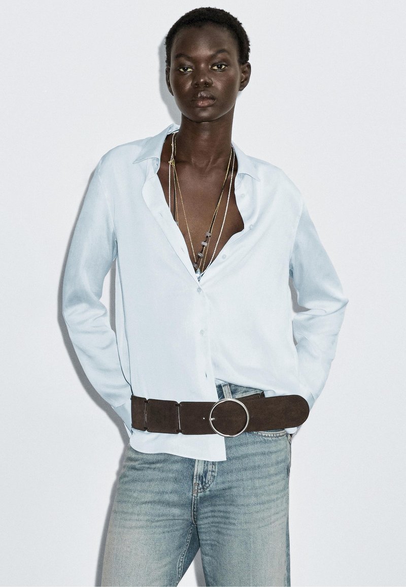 Light blue button-up shirt with long sleeves, paired with light blue denim jeans and a brown suede belt featuring a silver circular buckle.