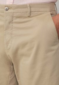 Beige Baumwollhose mit glatter Textur, ausgestattet mit einem einzigen Knopfverschluss und ordentlich gearbeiteten Taschen. Lässiges Design, leichtes Material.