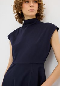 Mekko, jossa on laivastonsininen väri, hihaton muotoilu ja korkea kaulus. Materiaali on sileä ja istuva yläosa yhdistyy levenevään hameosaan. Mekossa on puhtaat linjat ja minimalistinen muotoilu.