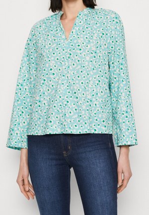 Blusa de manga larga azul claro con un patrón floral en blanco y verde; presenta un escote en V y un ajuste relajado. Combinada con jeans de mezclilla oscura.