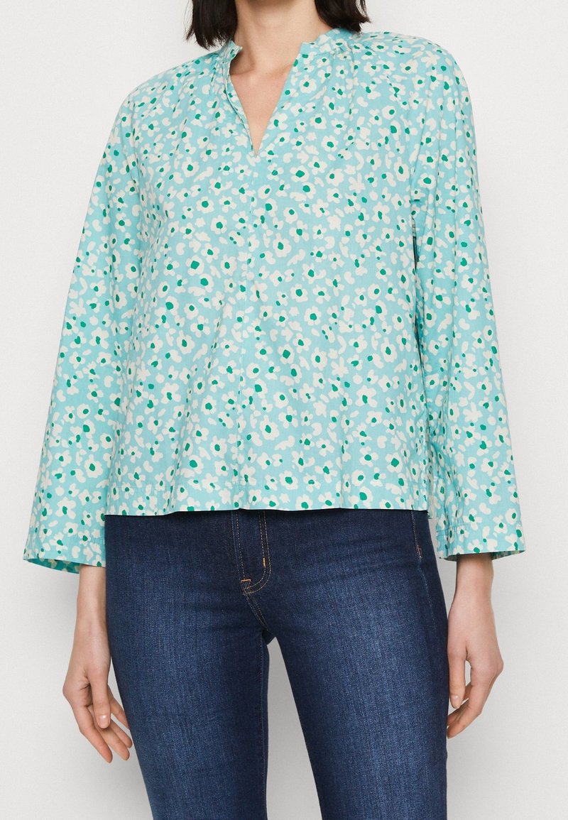 Ljust blå långärmad blus med ett blommönster i vitt och grönt; har V-ringning och avslappnad passform. Matchad med mörk denimjeans.