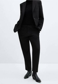 Person in einem schwarzen Blazer, passenden hochtaillierten Hosen, schwarzem Rollkragenpullover und schwarzen Absatzstiefeletten, der mit Händen in den Taschen dasteht.