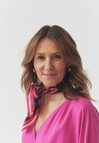 Pink blouse with a V-neck, paired with a patterned scarf featuring hearts in vibrant colors on a dark background. Features soft, wavy hair.