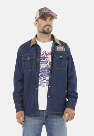 Homme portant une casquette beige et marine, une veste en denim bleu avec un col marron par-dessus un t-shirt blanc à motif et un jean bleu, debout devant un fond blanc.