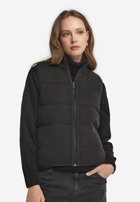 Svart pufferväst med en texturerad yta, dragkedjestängning och stickade detaljer, kombinerad med en svart polotröja och mörk denim.