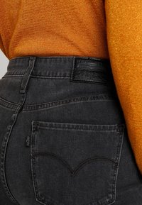 Jeans de mezclilla negra con un diseño distintivo en el bolsillo trasero que presenta una costura curva, combinados con un suéter naranja texturizado.