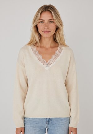 Donna bionda con un maglione crema a scollo a V con rifiniture in pizzo e jeans azzurri chiari, in piedi davanti a uno sfondo chiaro uniforme.