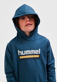Blauer Hoodie aus weichem Stoff, mit einem großen weißen und orangefarbenen Logo mit der Aufschrift "hummel SPORTSWEAR" über der Brust.