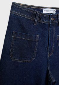 Tmavomodré denimové džínsy so žltým prešívaním, dizajn s piatimi vreckami a štandardným pásom s kovovým gombíkom a štítkom.