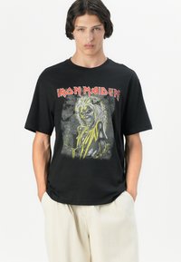 Melna kokvilnas t-krekls ar grafisku attēlu, kurā attēlots varonis ar gariem matiem un skeleta seju. Uz krekla ir izcils teksts "IRON MAIDEN" sarkanā krāsā.