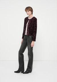 Jaunas žmogus, vilkintis bordiniame pailgintame švarkelyje su blizgučiais, šviesiai rožine marškinėliais, tamsiai pilkomis kelnėmis ir juodomis kulninėmis botais, stovi prieš paprastą baltą foną.