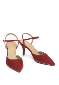 Červené semišové boty na vysokém podpatku s špičatou špičkou a nastavitelným kotníkovým páskem. Mají štíhlý podpatek a hladké vnitřní podšití.
