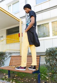 Junge Person trägt eine schwarze Mütze, ein grafisches T-Shirt, gelbe Hosen und gemusterte Turnschuhe und steht auf einer Holzbank im Freien in der Nähe von Wohnungsbriefkästen.