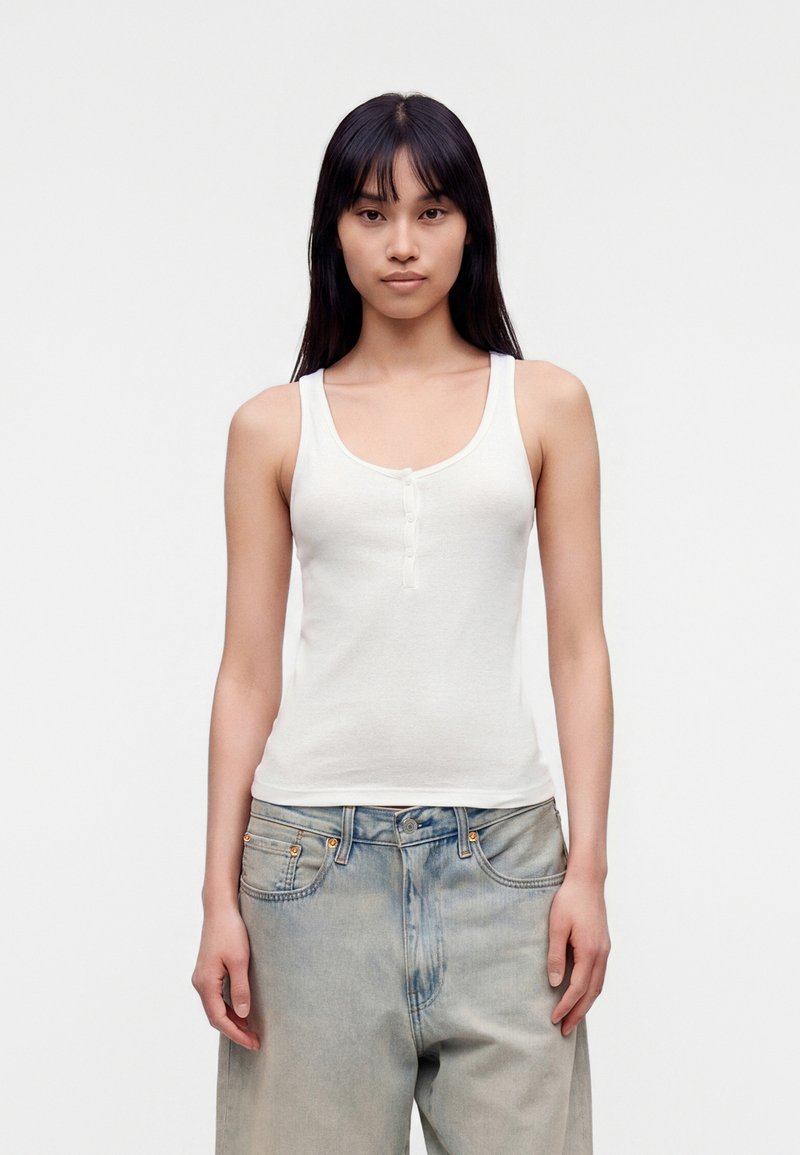 Femme aux longs cheveux foncés portant un débardeur blanc sans manches boutonné et un jean bleu clair ample, debout devant un fond uni.