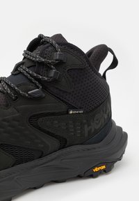 Fekete túracipő, texturált anyaggal, hálós részletekkel és Vibram talppal. GORE-TEX vízálló bevonattal és szürke fűzőkkel ellátva.