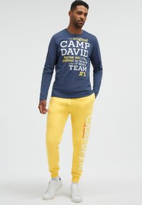 Mann trägt ein dunkelblaues Langarmhemd mit weißem und gelbem Text und leuchtend gelbe Jogginghose mit weißem und rotem Text, steht mit einer Hand in der Tasche.