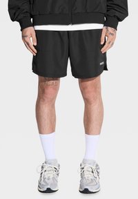 Czarne spodenki sportowe o luźnym kroju, z bocznymi kieszeniami i etykietą "PROSTO". Noszone z białymi skarpetkami do kolan i szarymi trampkami.