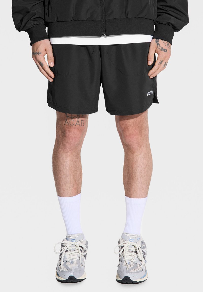 Czarne spodenki sportowe o luźnym kroju, z bocznymi kieszeniami i etykietą "PROSTO". Noszone z białymi skarpetkami do kolan i szarymi trampkami.