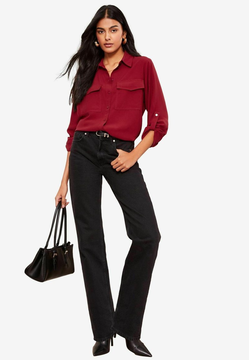 Chemise boutonnée bordeaux avec poches poitrine, associée à un jean noir à jambes droites. Le mannequin tient un sac à main noir et porte des escarpins à bout pointu.