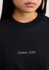 Camiseta de algodón negra con cuello redondo y mangas cortas; presenta el texto "Carhartt WIP" en azul claro en la parte delantera.