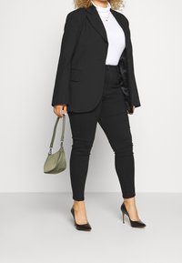 Svart kavaj över en vit polotröja, matchad med svarta skinny jeans och svarta högklackade skor. Håller en liten grön handväska.