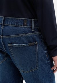 Ciemnoniebieskie jeansy denimowe o standardowym kroju, wyposażone w tylne kieszenie, żółte przeszycia i czarną metkę na pasku od spodni.