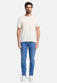 T-shirt beige clair à manches courtes en tissu texturé, associé à un jean slim bleu et des baskets blanches, porté par un homme debout.