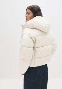 Młoda kobieta w kremowej pikowanej kurtce z kapturem, zwrócona w lewo, stojąca na jednolitym białym tle, ubrana w ciemnoniebieskie dżinsy.