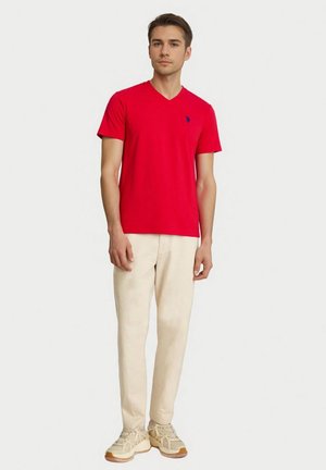 Rode katoenen v-hals T-shirt met korte mouwen, met een klein marineblauw logo op de borst. Draagt lichte broek en sneakers.