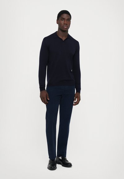 Man standing, wearing a dark navy long-sleeve polo shirt, dark blue trousers, and black leather shoes against a plain light background.