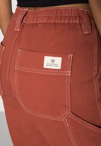 Kalhoty v rezavé barvě vyrobené z texturované tkaniny, s elastickým pasem a viditelným stehováním. Disponují zadní kapsou s bílou etiketou označující styl.