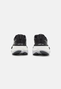 Svarta sportskor med en texturerad ovansida, kontrasterande vit sula och förstärkt häl för stabilitet. Noterbar logotypdetalj på sidan.