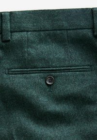 Green wool trousers with a textured finish, featuring a back pocket secured by a black button and tailored stitching accents.
