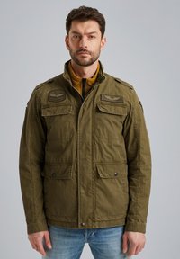 Veste olive verte avec fermeture éclair à l'avant et boutons pression, deux poches poitrine, épaulettes et doublure jaune sous le col.