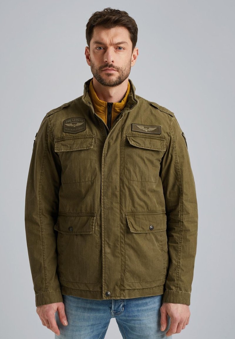 Veste olive verte avec fermeture éclair à l'avant et boutons pression, deux poches poitrine, épaulettes et doublure jaune sous le col.